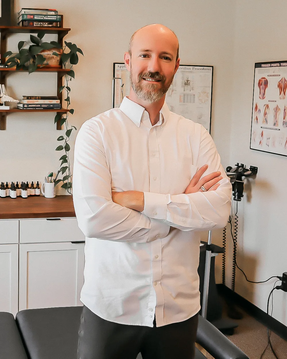 Dr. Andrew Rostenberg standing in his treatment room at Red Mountain Natural Medicine in Boise, Idaho — chiropractic table, anatomy charts, and supplement workspace behind him